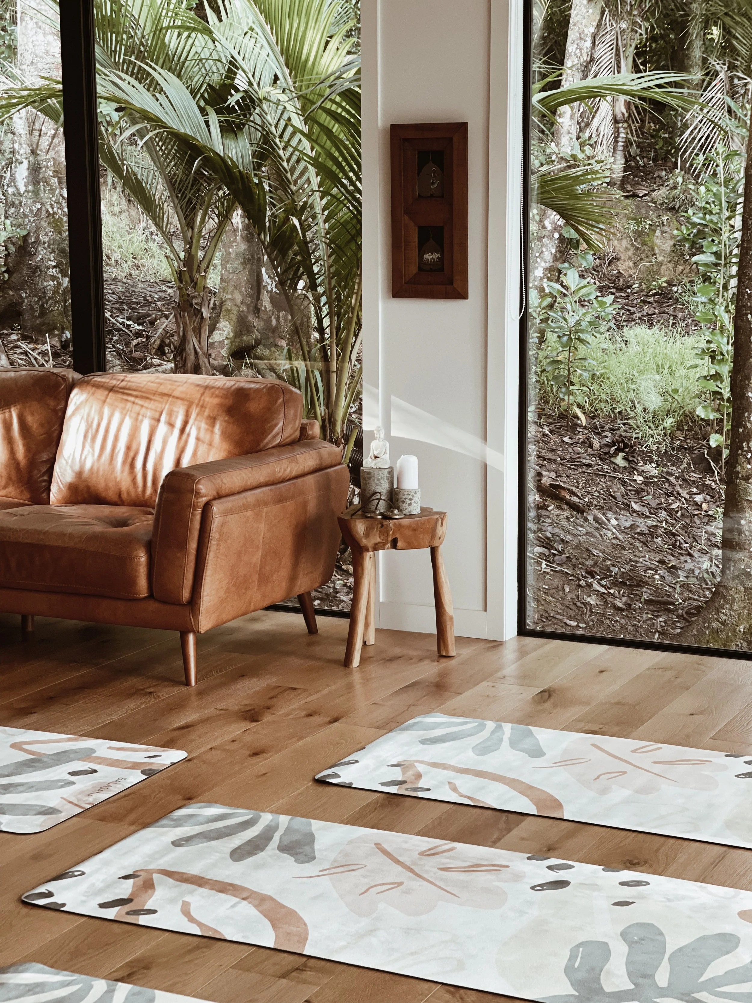 Interior space with a leather couch, decorative mats, wooden side table, and large windows showing a garden view.