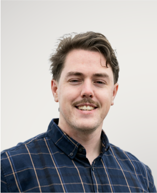 A young man with brown hair and a light mustache smiling at the camera, wearing a blue plaid shirt, with snow-capped mountains in the background.