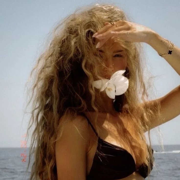 A woman with long, curly blonde hair standing on a beach, holding her hand over her forehead, with a white flower in her mouth, wearing a black bikini.