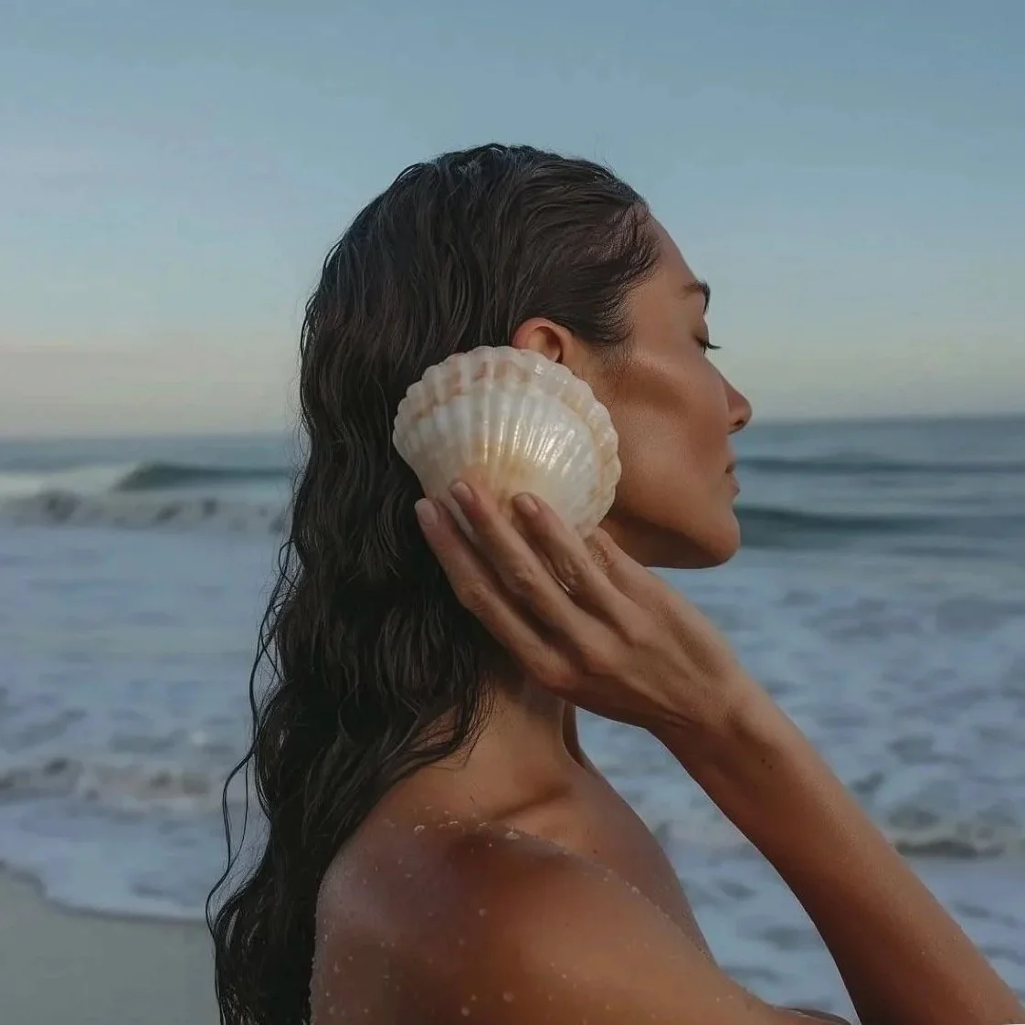 A woman with wet hair at the beach holding a seashell near her ear with her eyes closed.