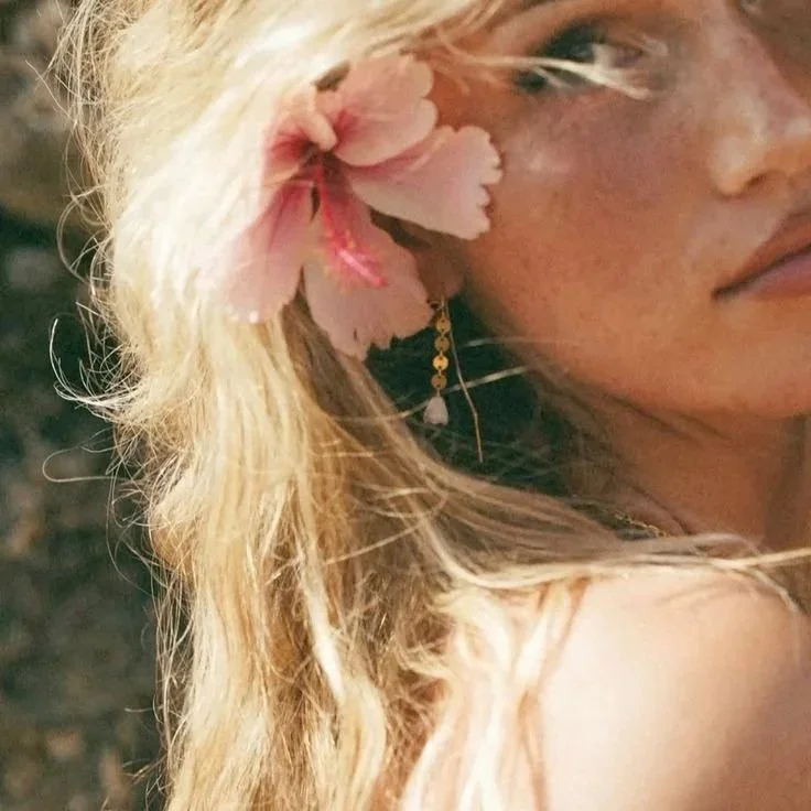 Close-up of a woman with blonde hair, wearing a pink flower hair accessory and a gold earring, showing part of her face and shoulder.