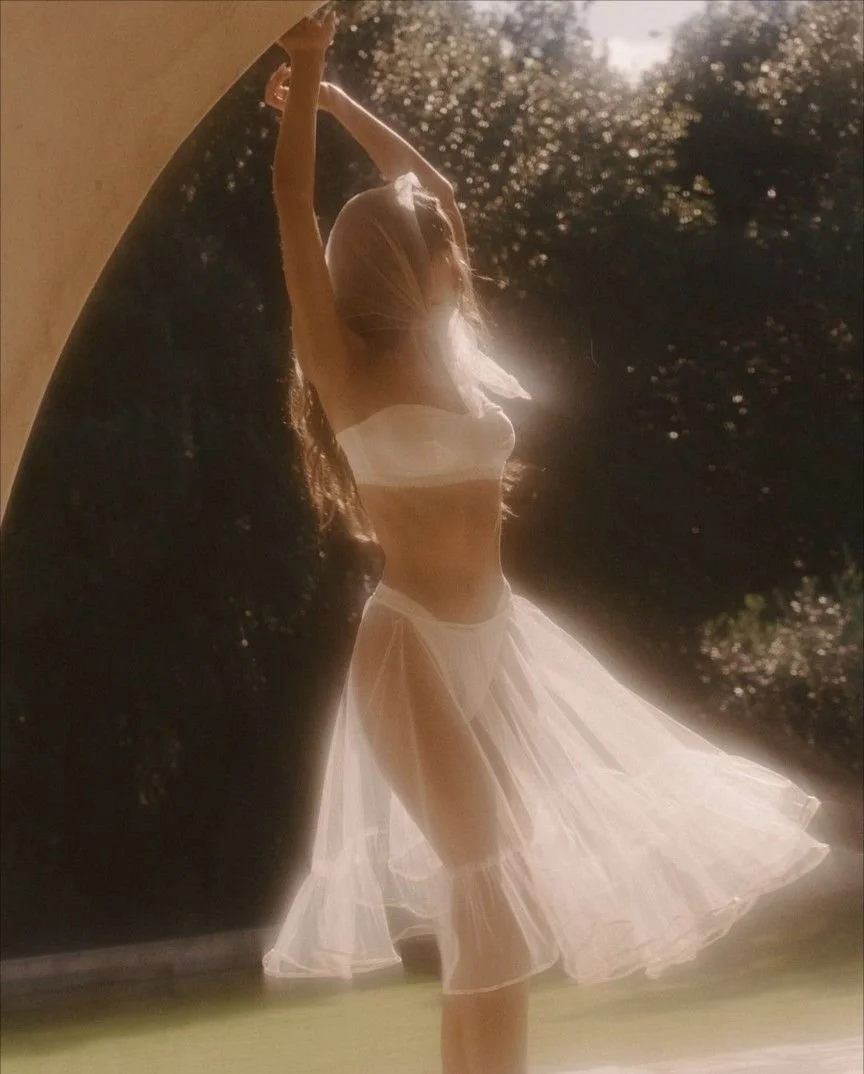 A woman in a flowing white dress stands under a bridge, with sunlight filtering through the leaves behind her, creating a dreamy atmosphere.