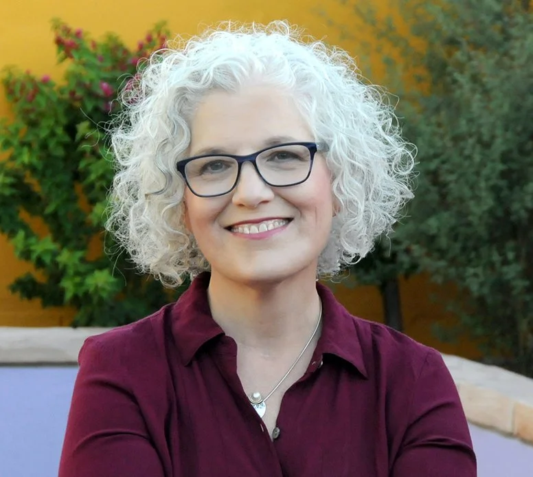 A woman with curly gray hair, glasses, and a maroon shirt outdoors, smiling in front of greenery and a yellow wall.
