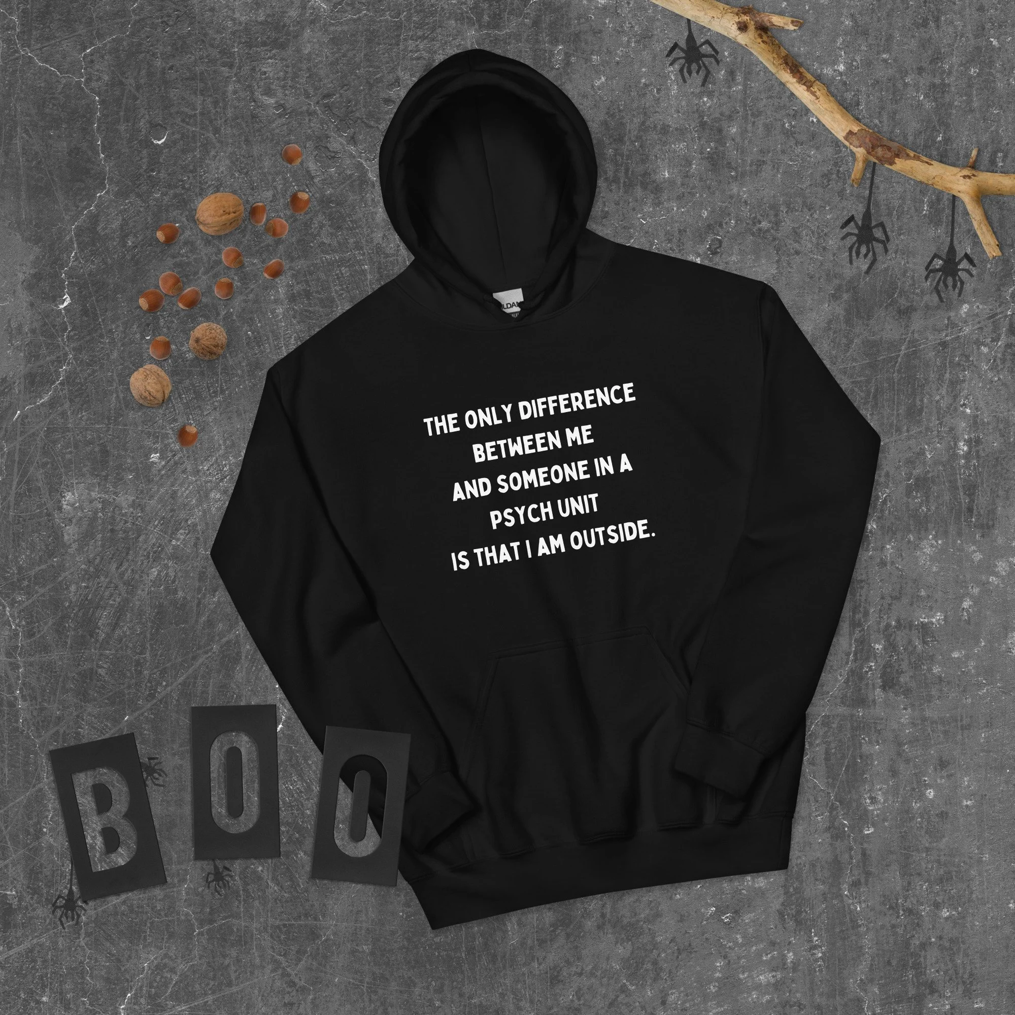 Black hoodie with white text on it, surrounded by a branch with hanging spider decorations, scattered nuts, and the word "BOO" spelled out on black cards, on a gray textured surface.