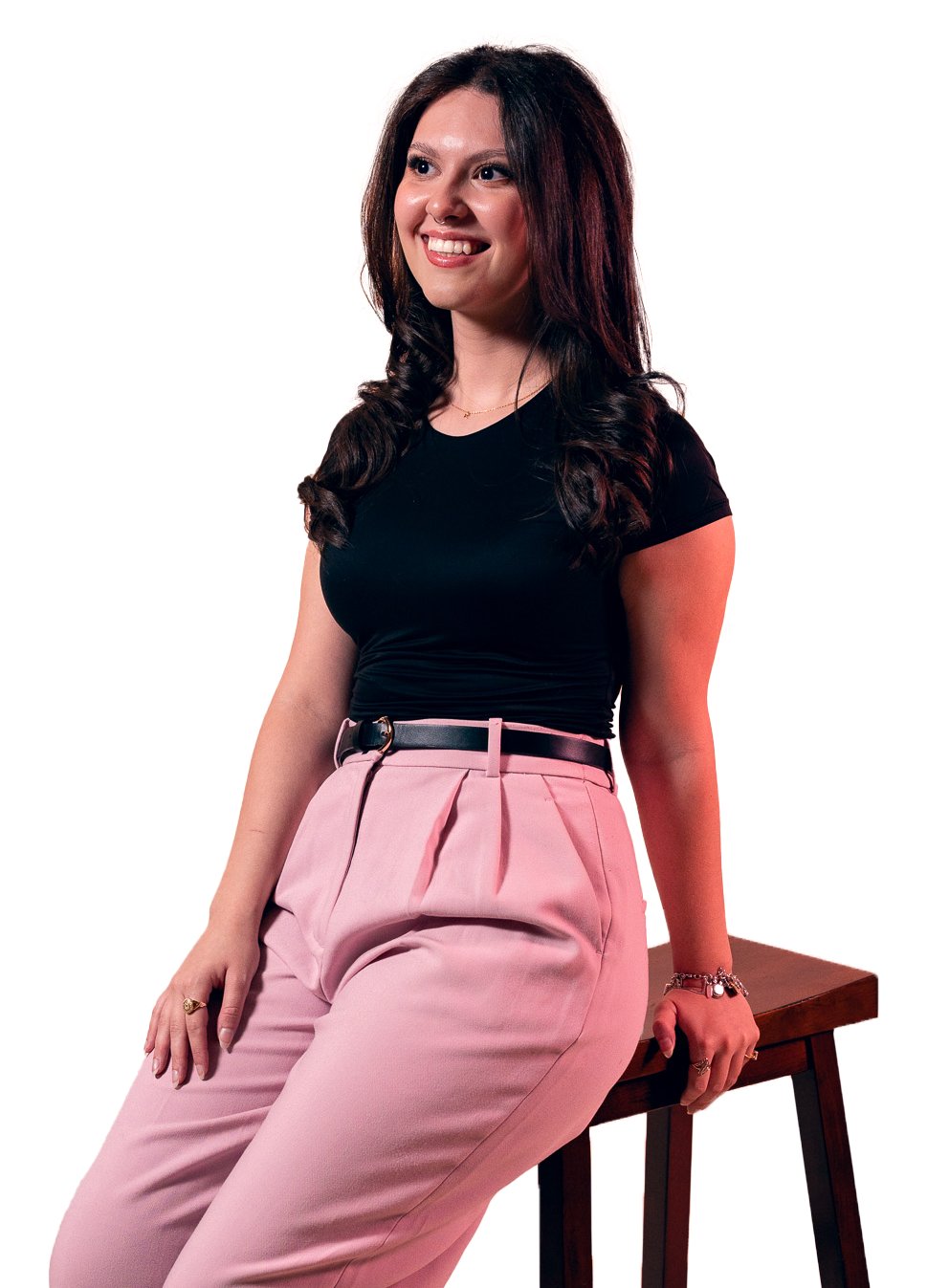 A young woman with long dark curly hair smiling, wearing a black t-shirt, pink high-waisted pants, and a black belt, sitting on a wooden stool.