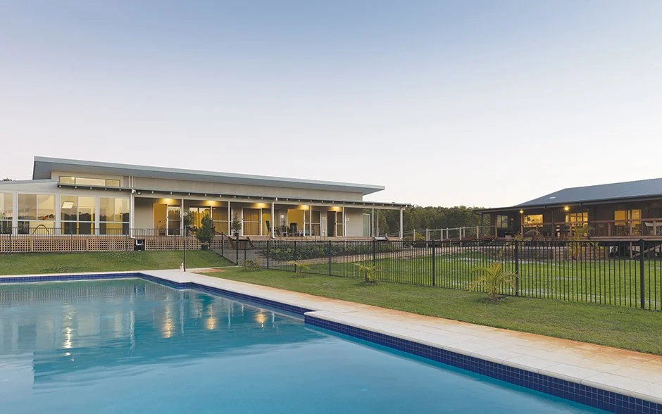 Residential backyard with a swimming pool, a modern house with large windows and a covered patio, and another house with a deck, all surrounded by a black fence and green grass.
