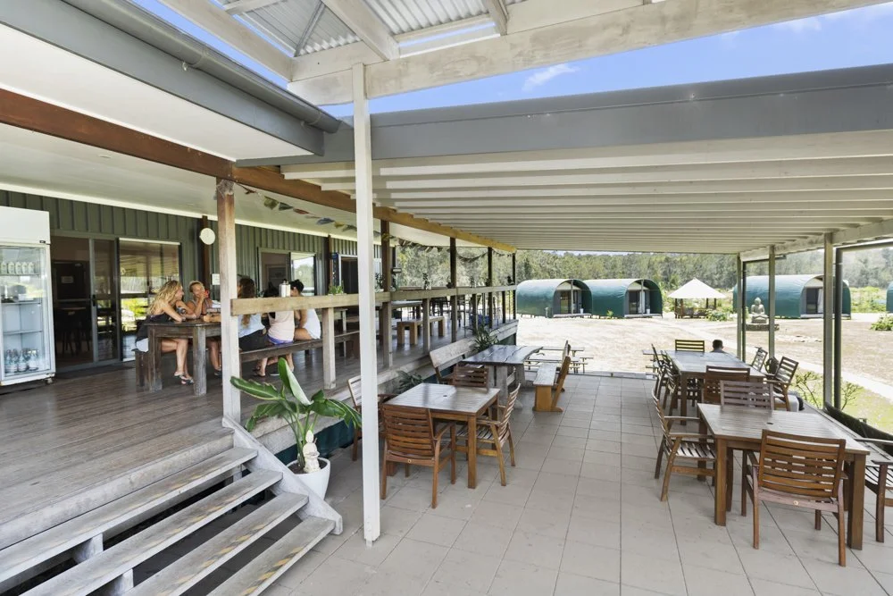 Outdoor dining area at a restaurant with wooden tables and chairs, some people sitting and talking, and green igloos in the background.