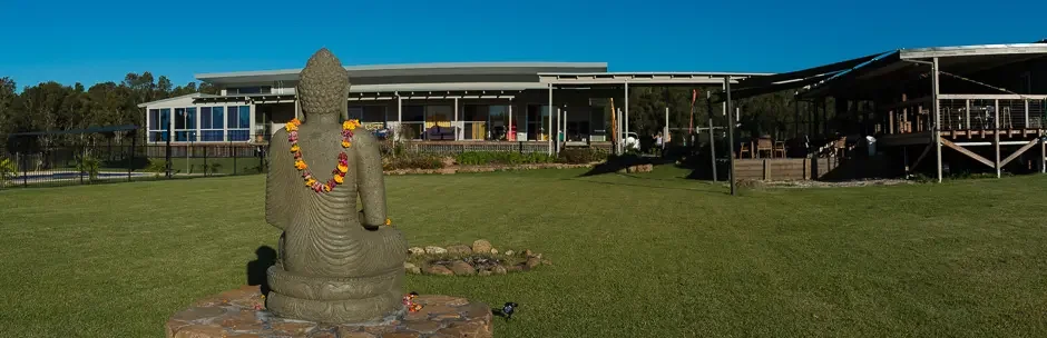 Stone Buddha statue with a flower garland sitting outdoors on grass in front of a modern building under a clear blue sky.