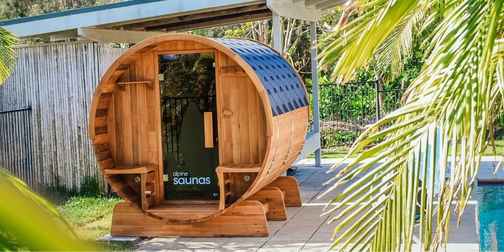 Wood-fired outdoor sauna in a backyard with a pool and tropical plants.