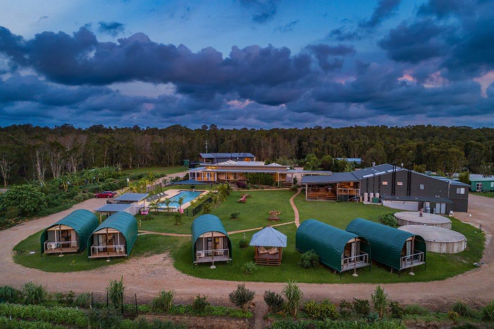 Aerial view of a resort with several green tent cabins, a swimming pool, a lounge area with umbrellas, and modern buildings, surrounded by trees and farmland under a cloudy sky.