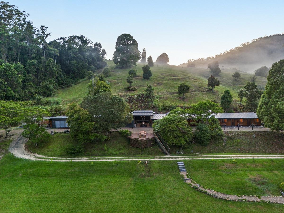 A hillside property with a house or barn, surrounded by trees and grassy slopes, with a winding path leading to it and mist in the background.