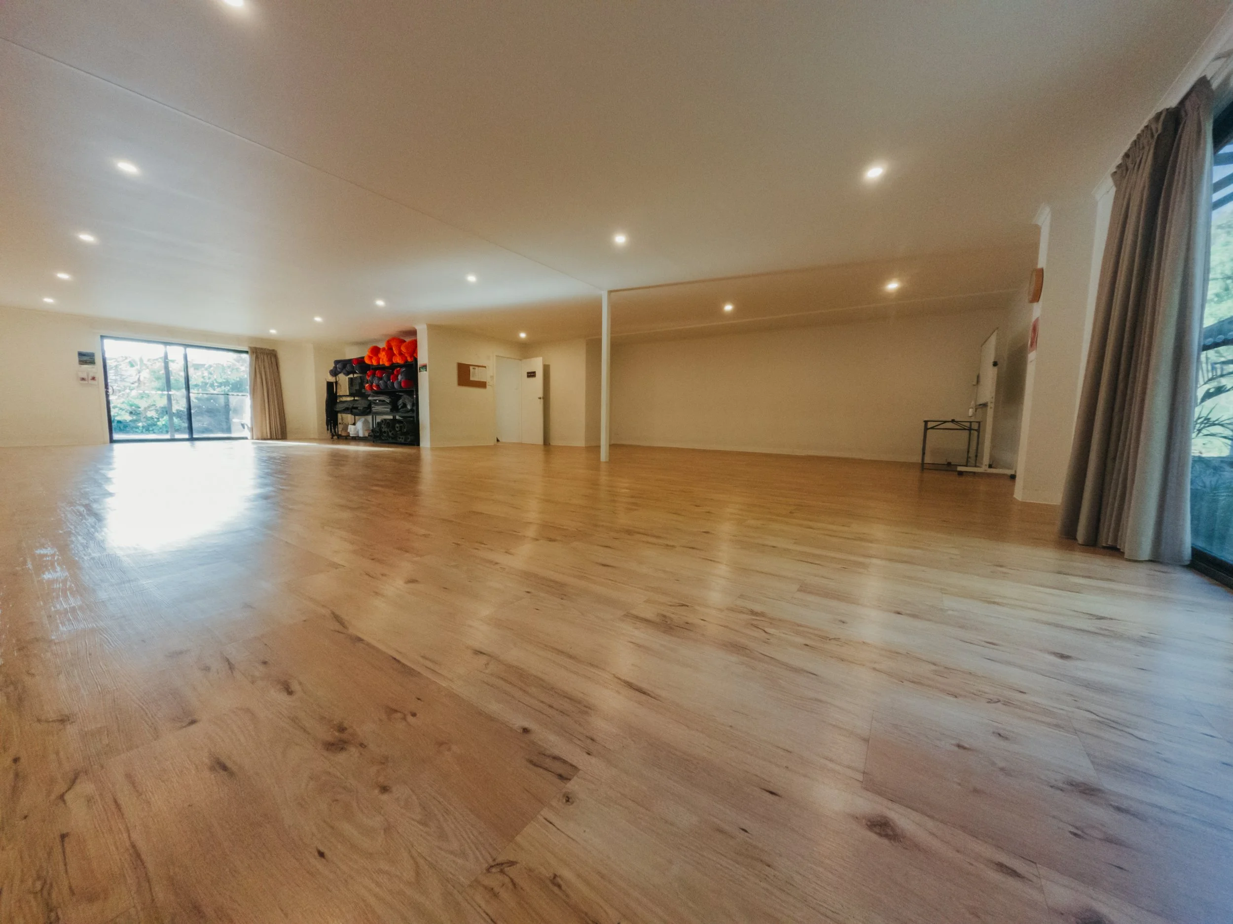 Empty indoor room with wooden flooring, large windows with curtains, and fitness equipment stored on shelves.