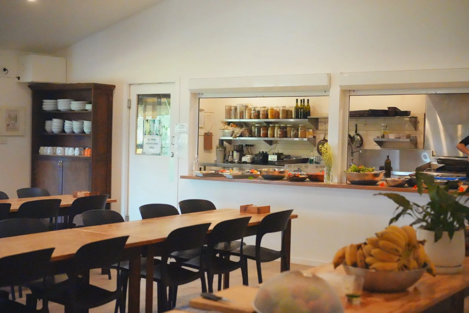 Inside a restaurant kitchen with a serving window, metal shelves with spices and bottles, and a dining area with wooden tables and black chairs, decorated with yellow flowers and bananas.