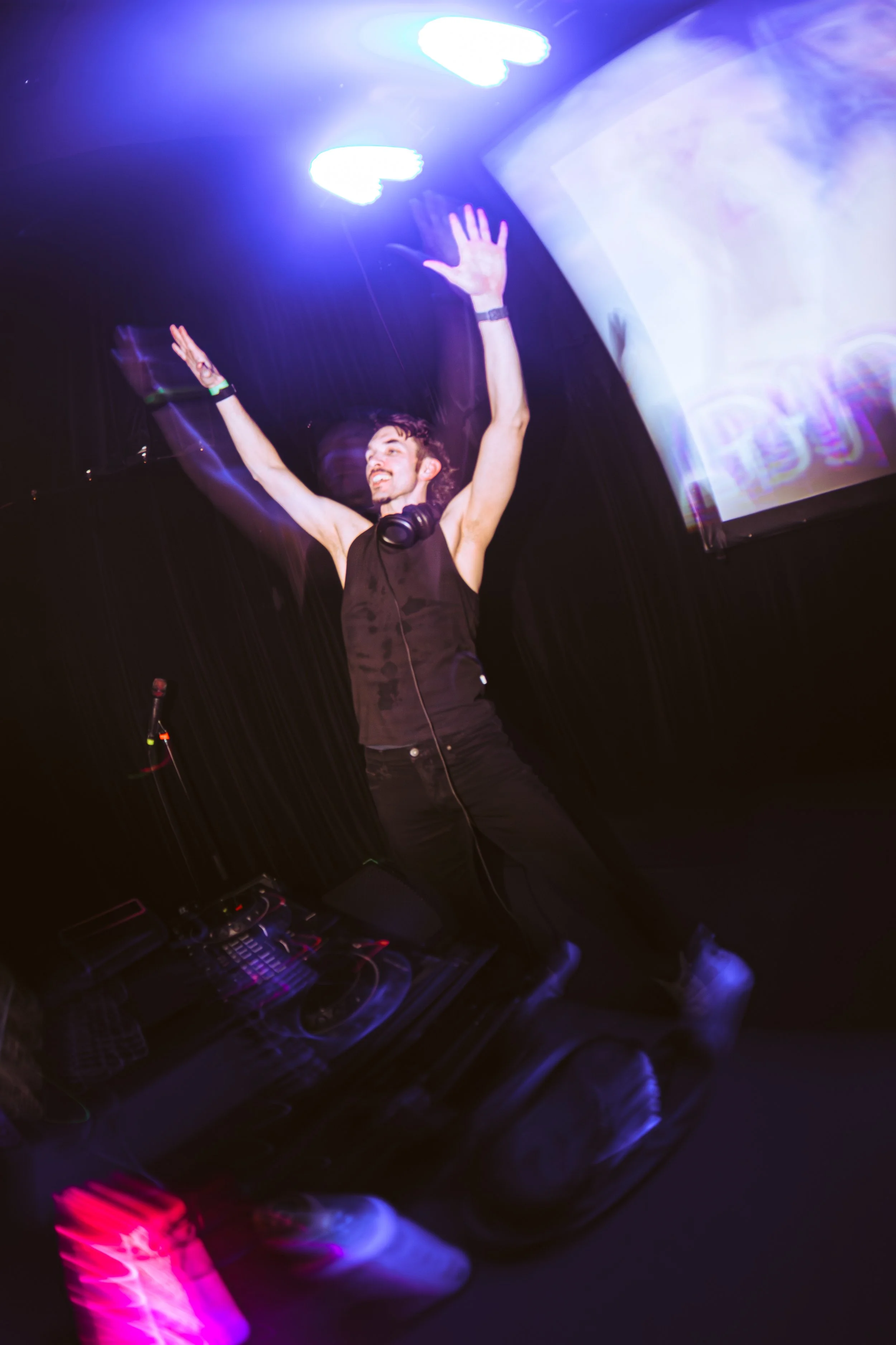 A male DJ is dancing with his arms raised in a dark club with colorful lights and a large screen in the background.