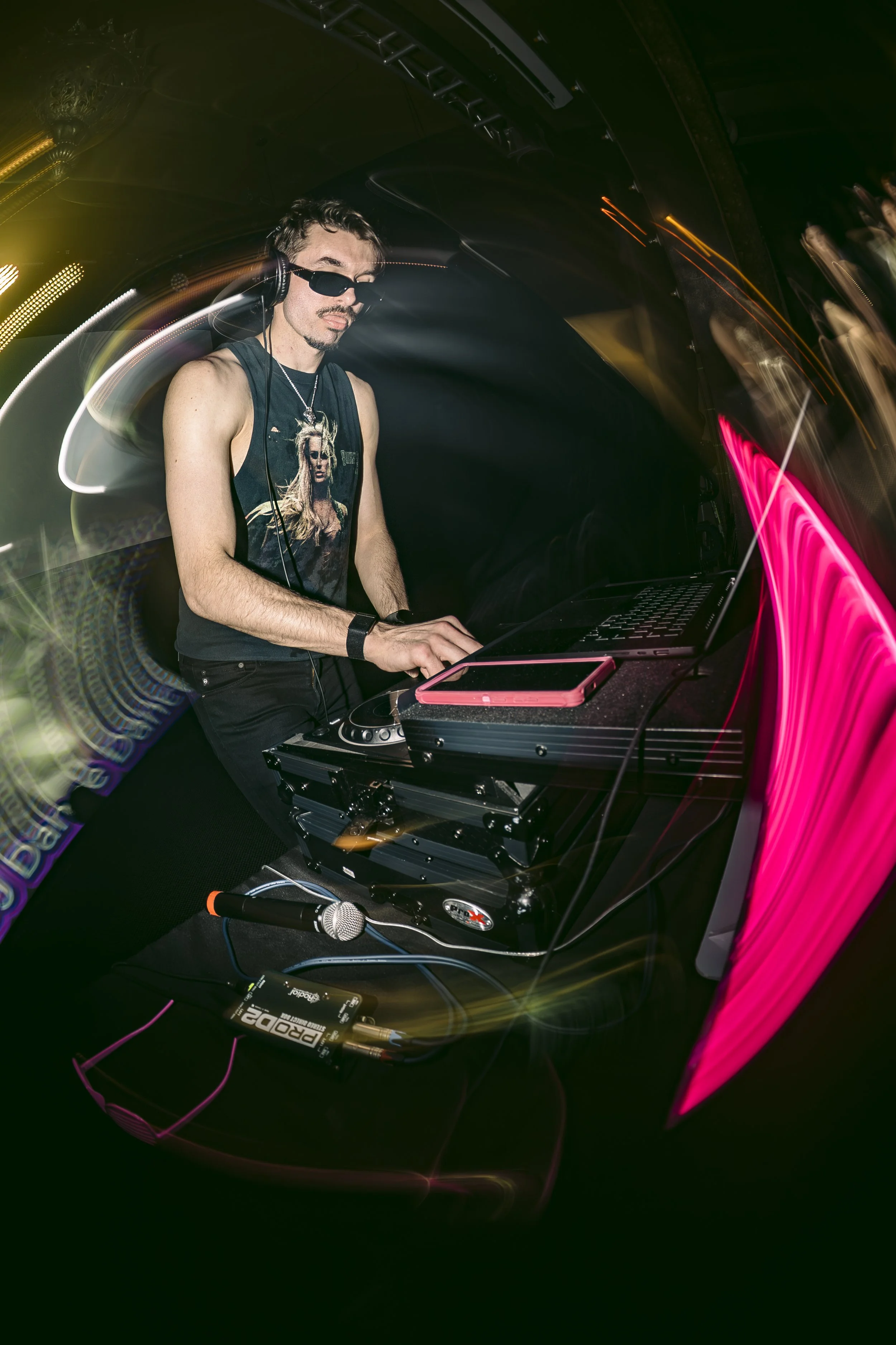 DJ wearing sunglasses, headphones, and a sleeveless shirt, performing at a nightclub with colorful lights and a laptop on a table.