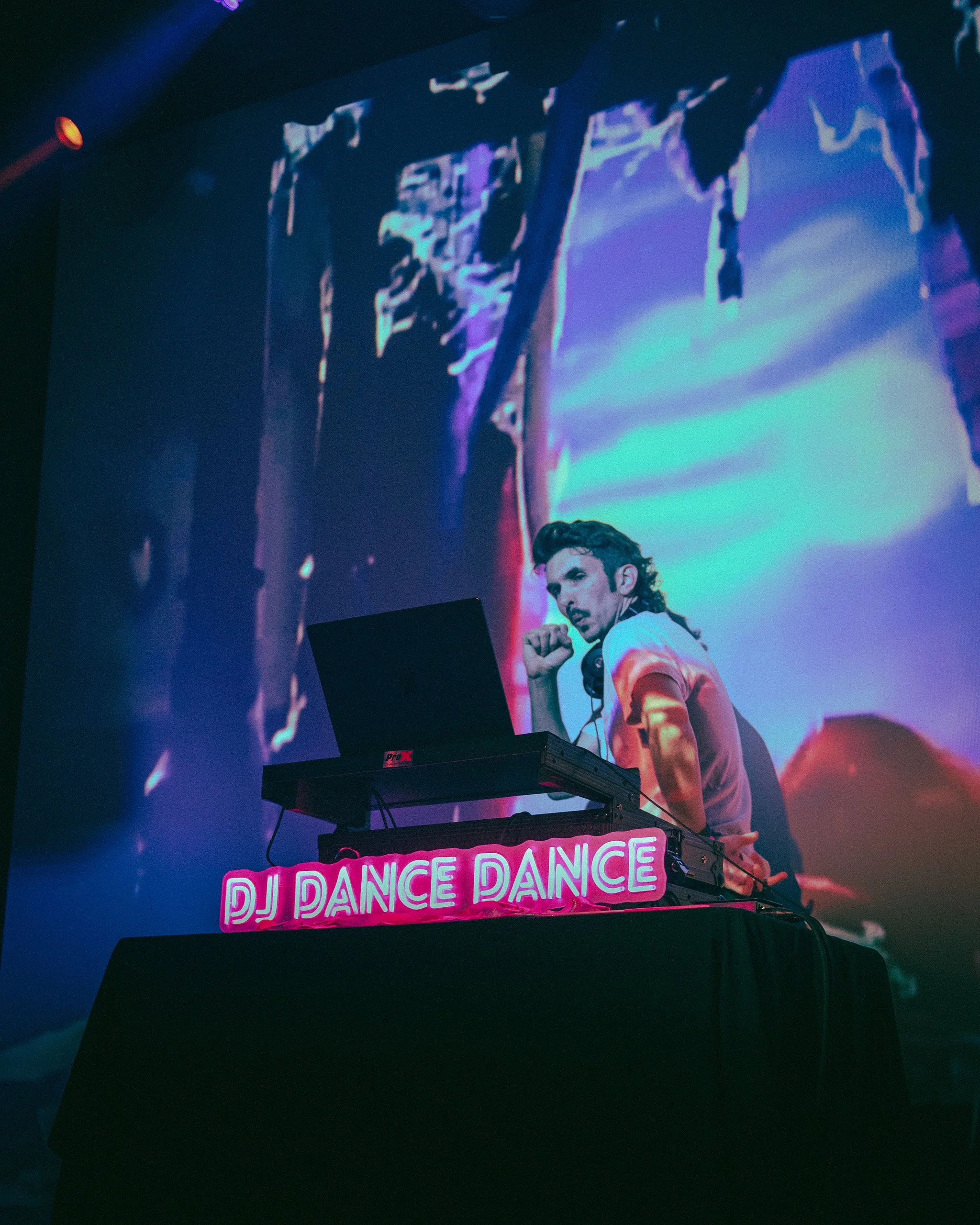 A DJ is performing at a nightclub, with a colorful digital backdrop behind him. There is a pink neon sign on the DJ booth that reads 'DJ DANCE DANCE'.