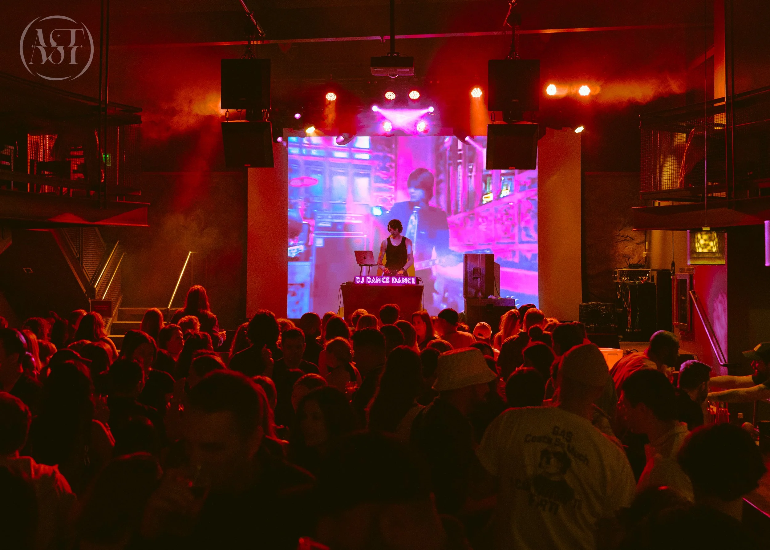 A DJ performing at a nightclub with a large crowd of people dancing. The DJ is on stage with a digital screen behind her displaying colorful visuals. The lighting is vibrant and red, creating an energetic atmosphere.