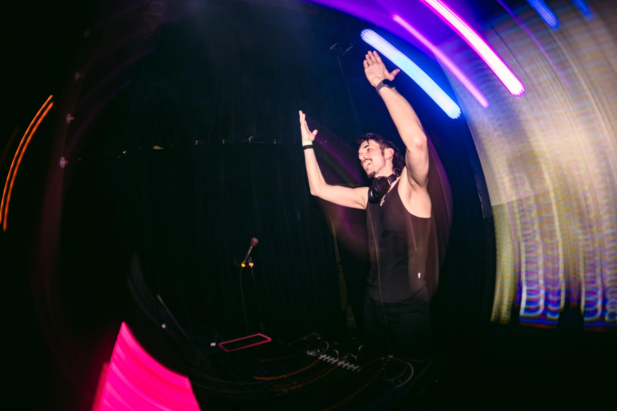 A DJ in a black sleeveless shirt with headphones around his neck, raising his hands with a joyful expression in a dark, colorful nightclub with neon lights.