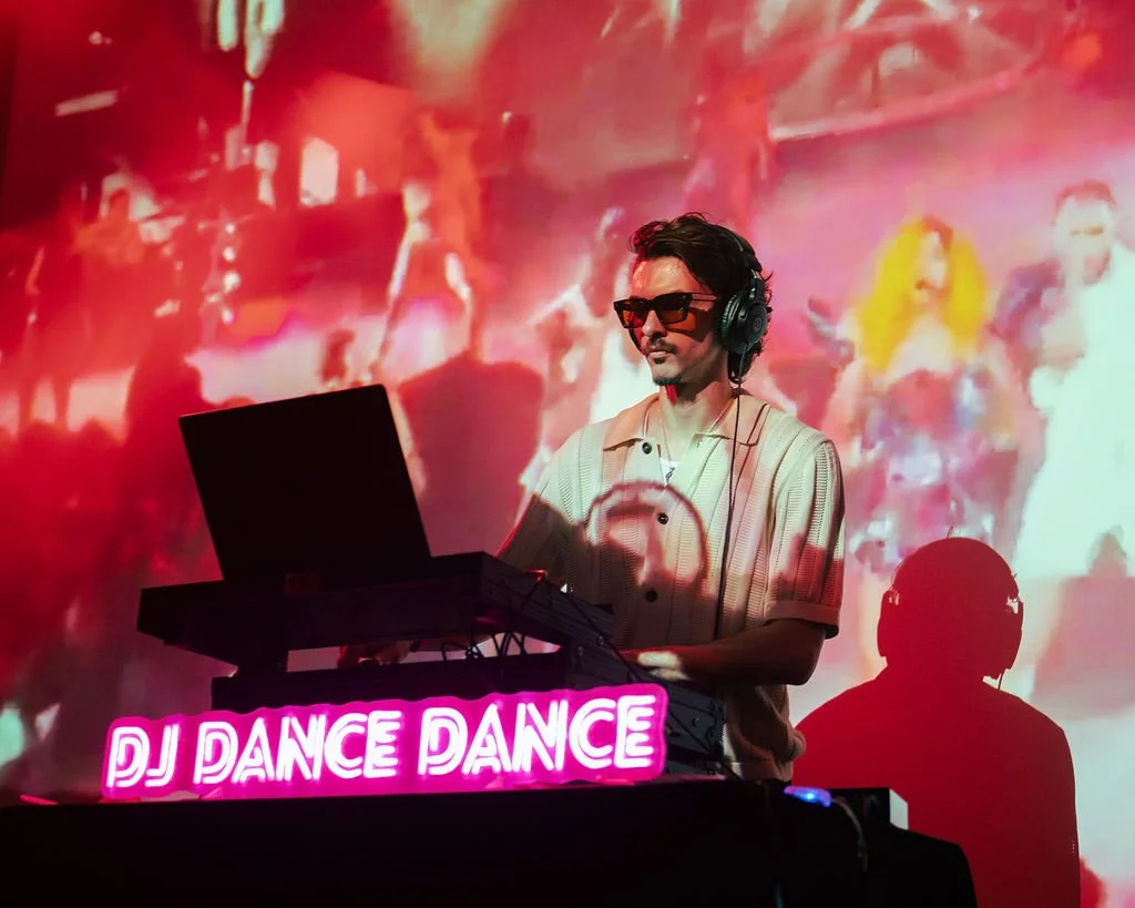 A DJ with sunglasses and headphones working on a music setup, with a neon sign reading 'DJ DANCE DANCE' and abstract colorful background images projected on the wall.