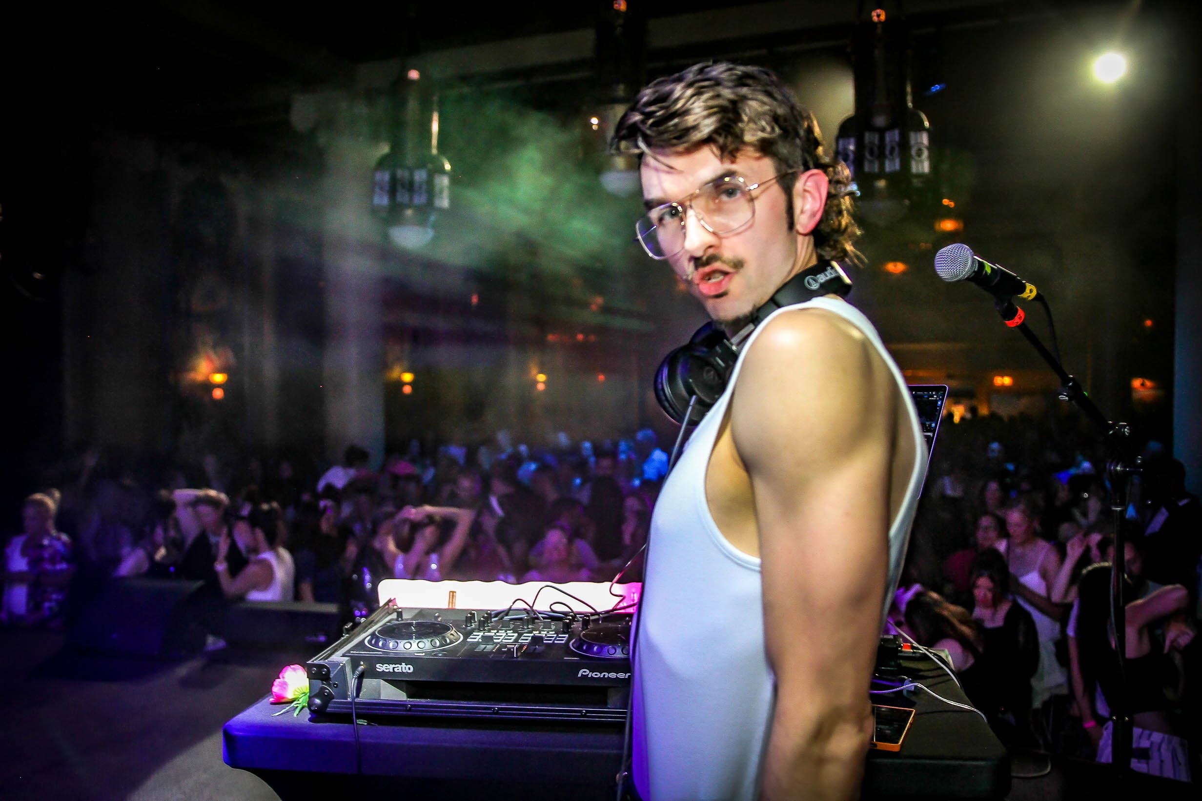 A DJ with glasses and a mustache wearing a white tank top, standing at a DJ booth with Pioneer and Serato equipment, on a stage with a microphone, with a crowd dancing in the dark, colorful club lighting, and fog effects in the background.