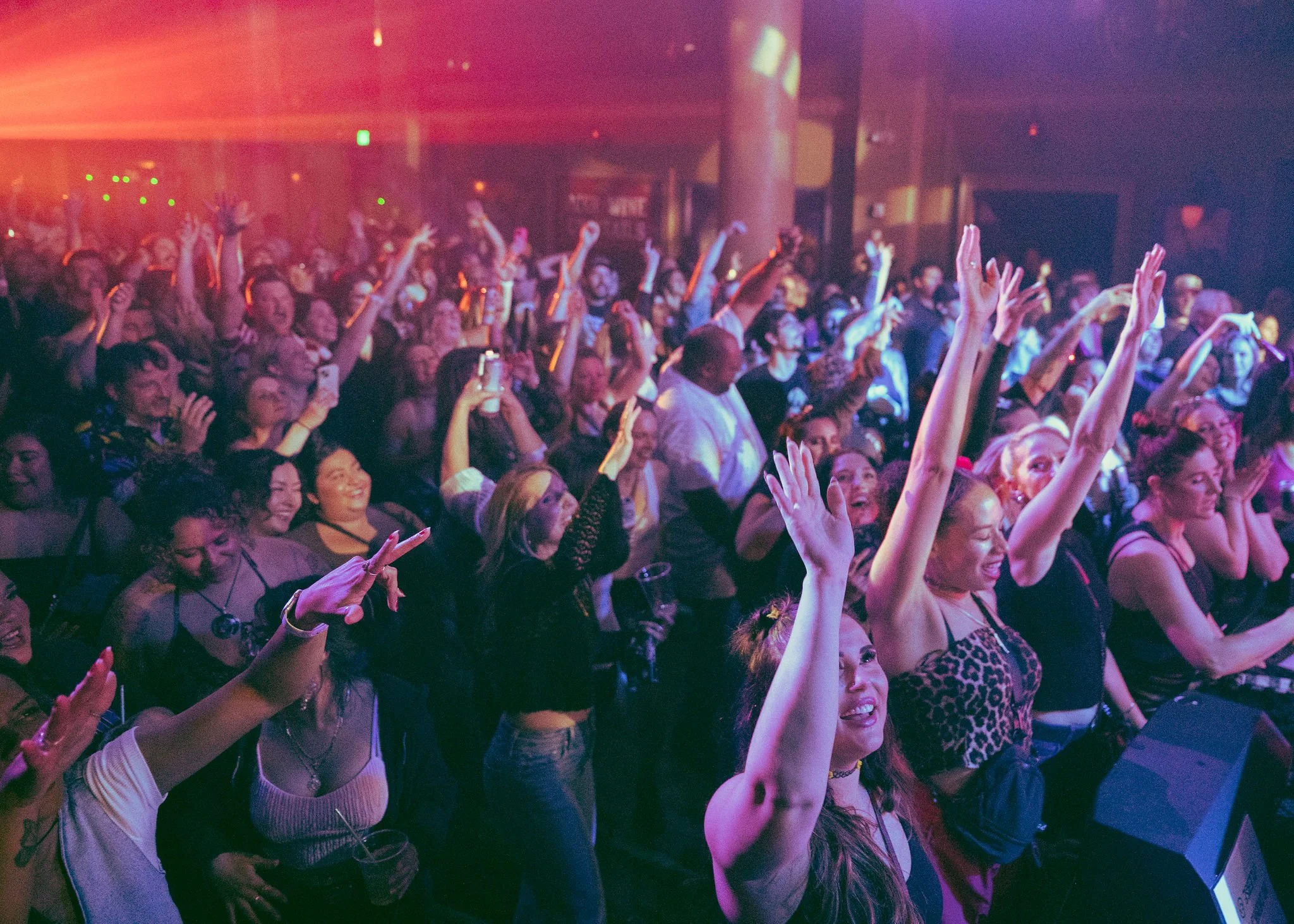 Crowd of people at a concert or event with many raising their hands, smiling, and enjoying the performance in a dimly lit space with colorful lights.