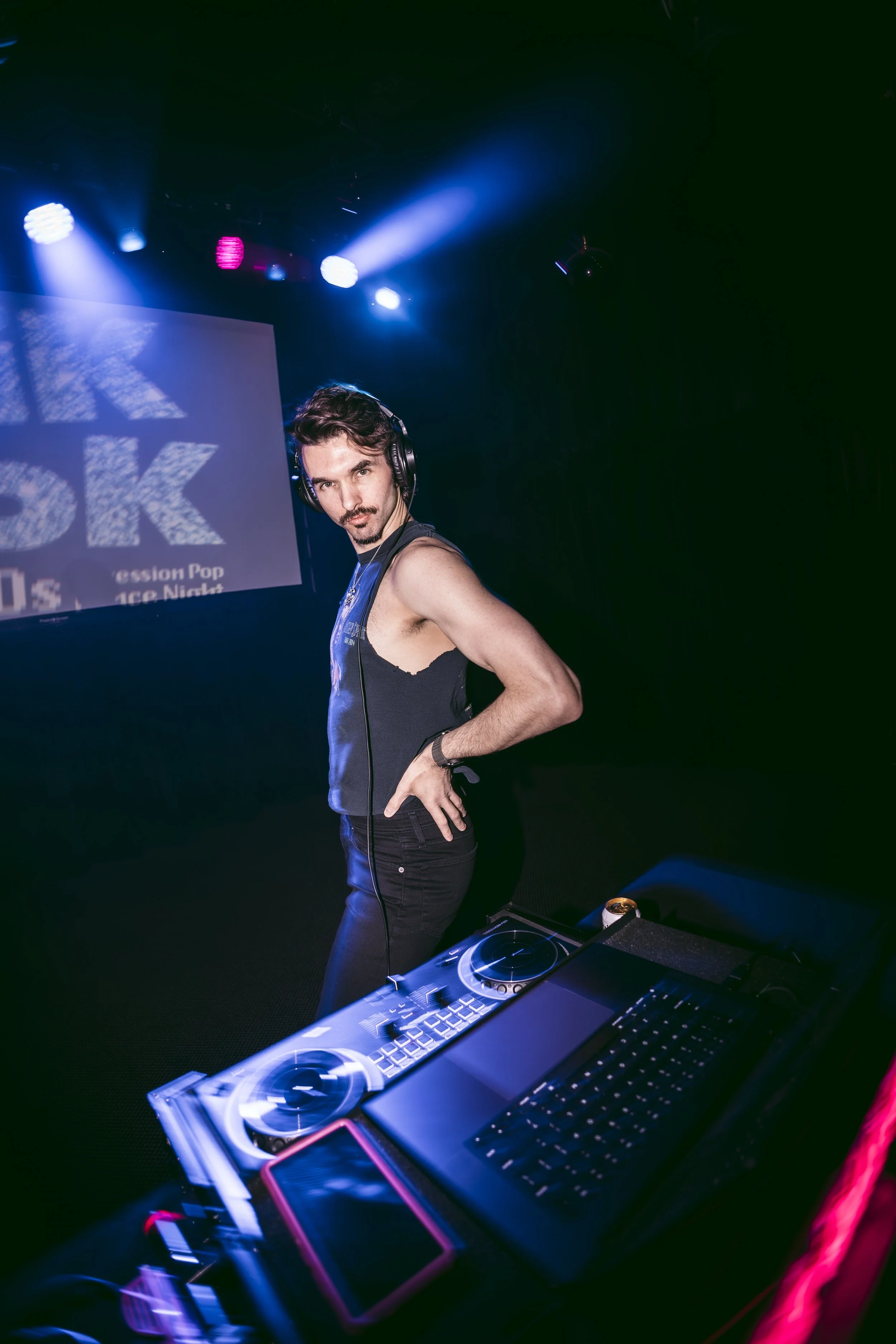 A young male DJ with dark hair and facial hair, wearing headphones and a sleeveless shirt, standing behind a DJ mixer and keyboard on a stage illuminated with blue and pink lights.