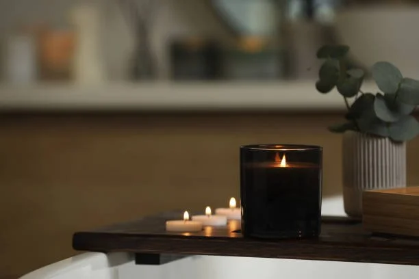 A lit candle in a black glass holder on a dark wooden surface, with several small unlit tealight candles nearby, and a potted plant with round leaves in a white ribbed pot in the background.