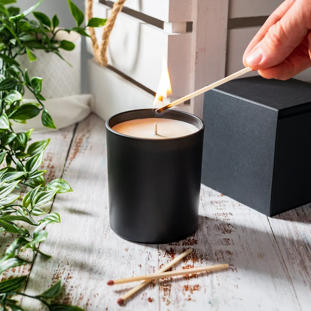 Person lighting a candle with a matchstick on a white wooden table, surrounded by greenery and a black box.