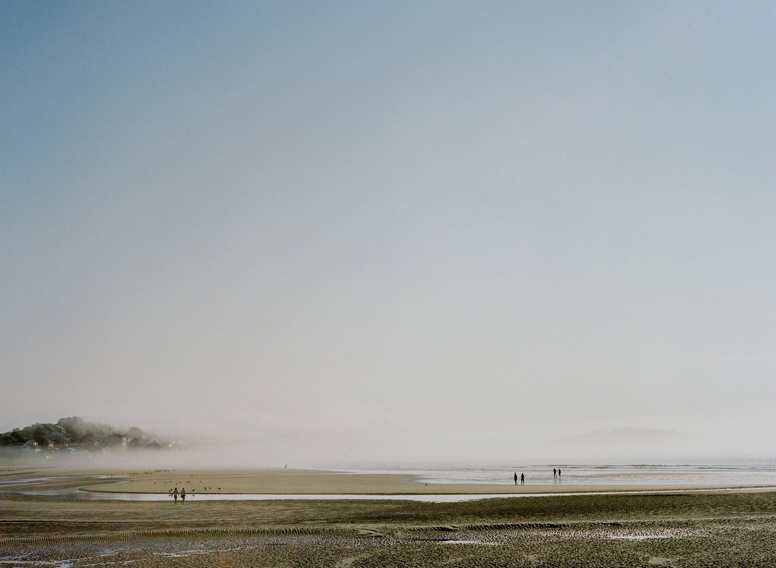 A foggy beach with a few people walking along the shore, low tide exposing wet sand and shallow water, with houses on the distant hillside partially obscured by fog.