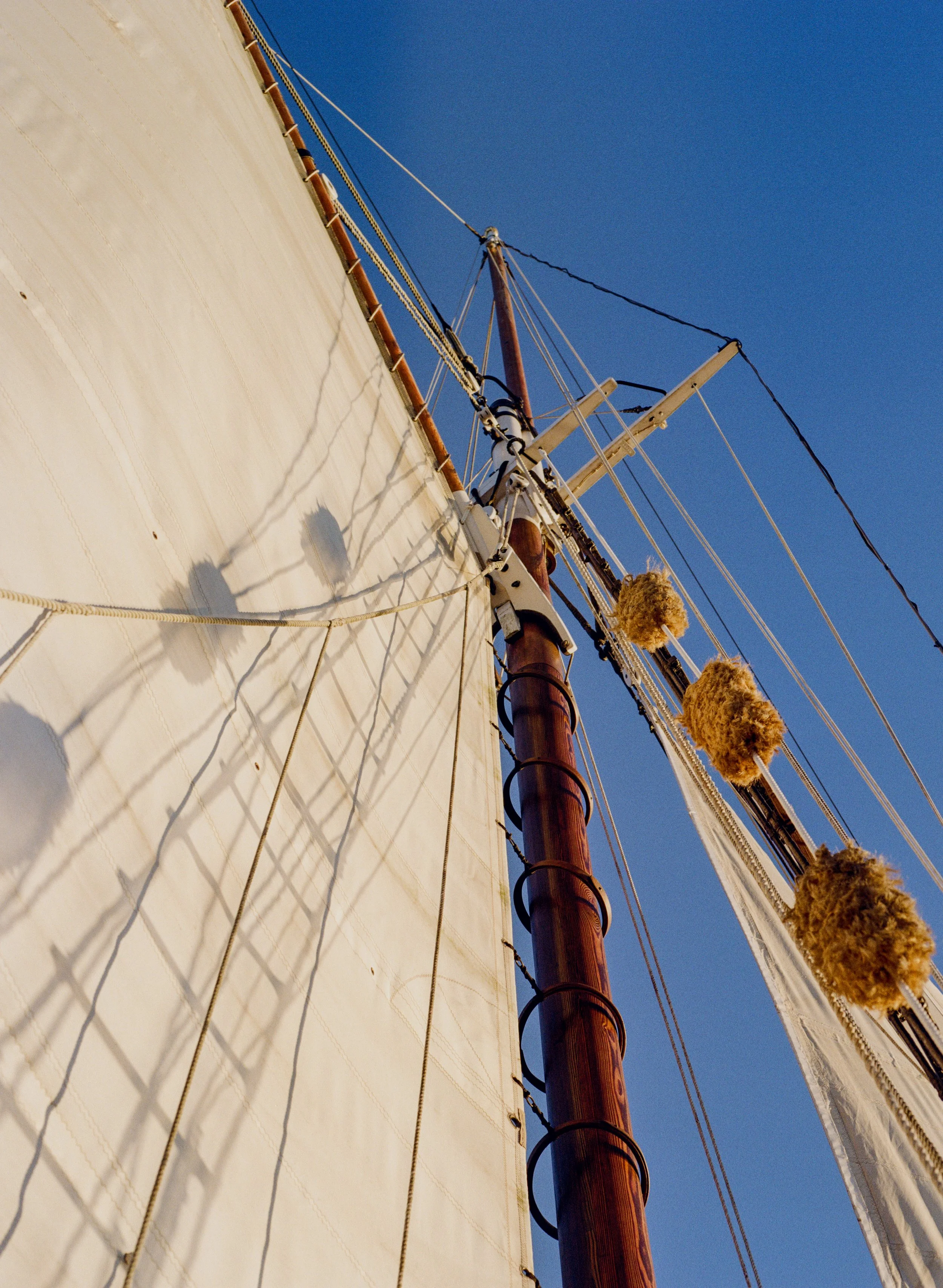 Upward view of a sailing ship's mast, rigging, and sails against a clear blue sky, with shadows of the rigging cast on the sail.