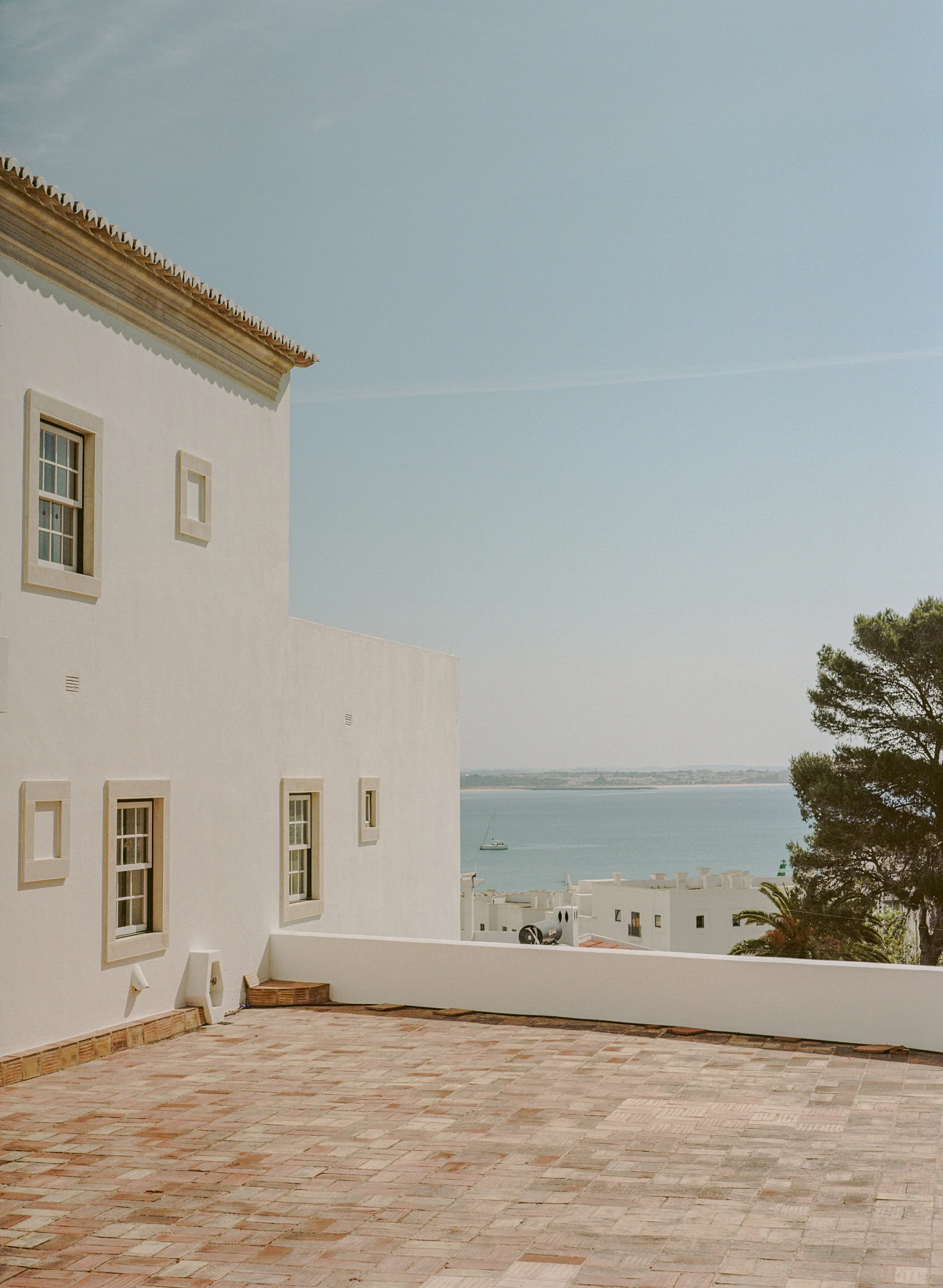 A white building with multiple small windows overlooking a body of water with a boat sailing. There are trees on the right and a brick-paved area in the foreground.