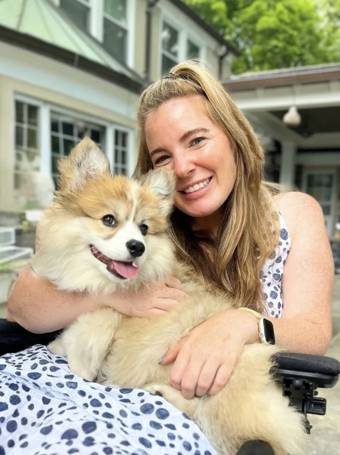 A woman smiling and holding a fluffy Corgi puppy outdoors on a patio with a house and trees in the background.