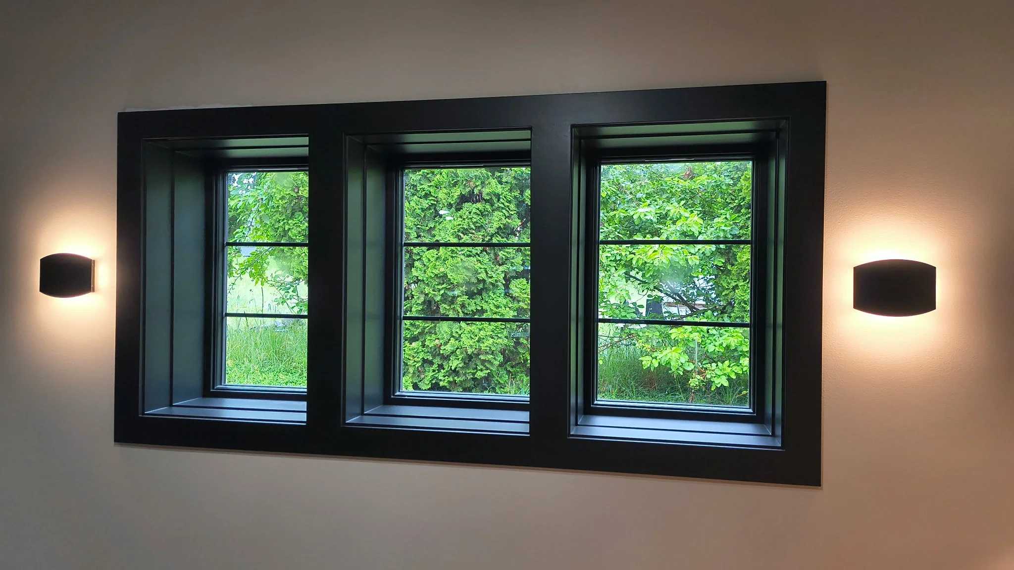 A multi-pane window with black trim on a white wall, showing green foliage outside, with two black wall-mounted lights flanking the window.
