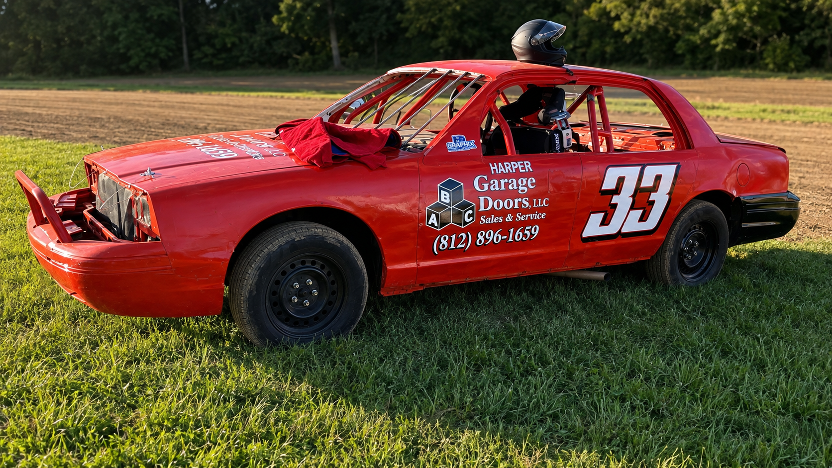 Red race car with the number 33 on the side, parked on grass, with a helmet on top and a racing harness inside, sponsored by Harper Garage Doors LLC.