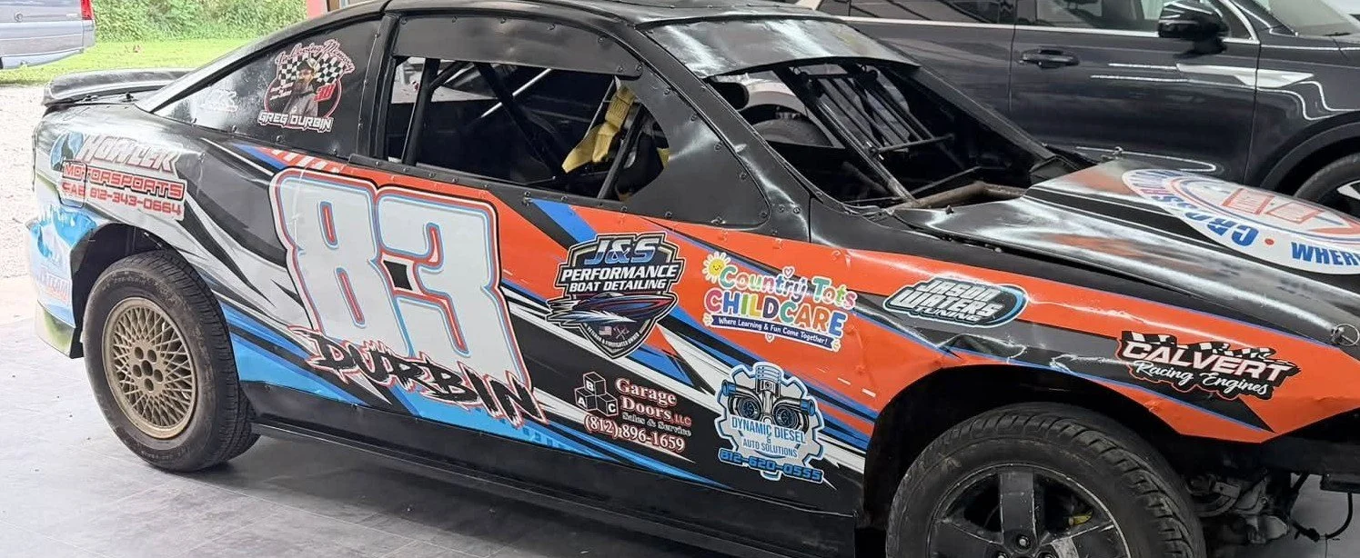 A race car with the number 83 on its side, decorated with sponsor decals and a black, orange, white, and blue color scheme, parked indoors next to other vehicles.