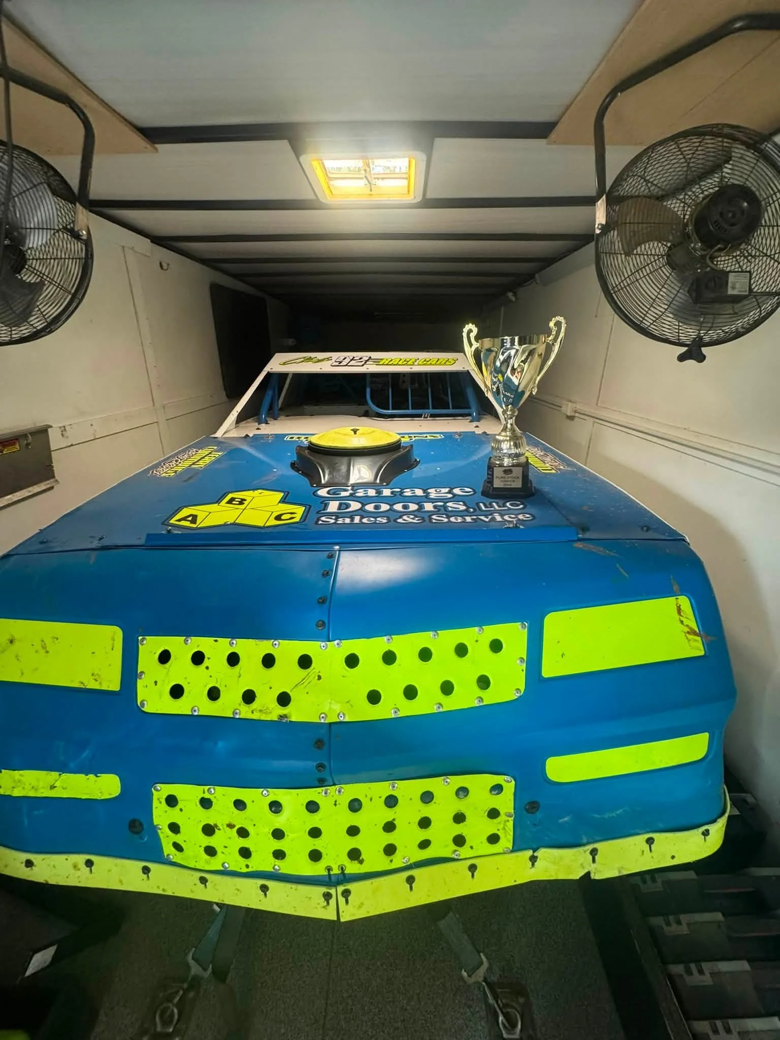 View from the front of a race car with blue and yellow decals, a trophy and a yellow air intake on the hood, inside a trailer or garage with fans and tables.