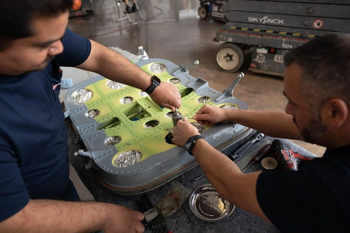 Two people working together on a mechanical or engineering project, using tools on a large, circular green and silver device with multiple circular openings, possibly part of an aircraft or aerospace component, in a workshop or industrial setting.