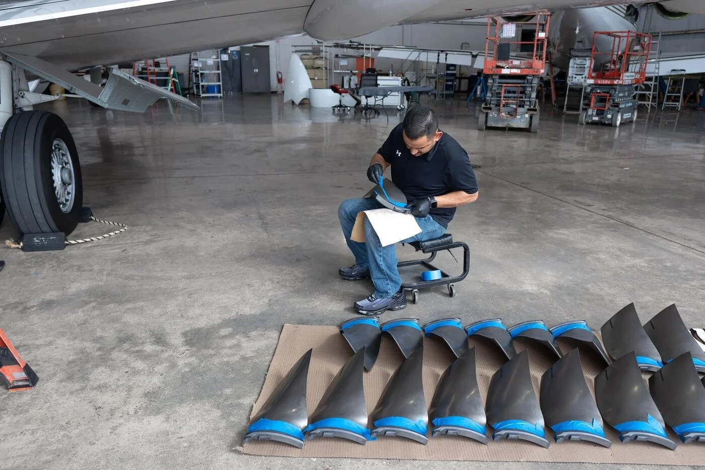 A worker in a warehouse or hangar is working on parts of an aircraft, possibly a jet, with various tools and equipment around him. The worker is seated on a small stool, examining or working on a piece. There are aircraft parts and work platforms visible in the background.