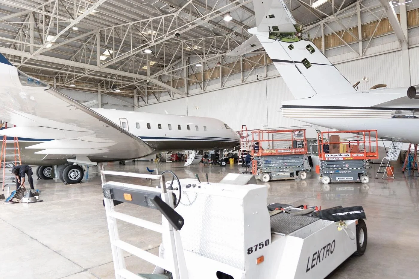 Inside an aircraft hangar where two private jets are undergoing maintenance or repair. Several technicians are working on the planes, and there is equipment and tools around. One jet's nose is visible on the left, and the tail of another jet is seen on the right with some maintenance personnel working nearby.