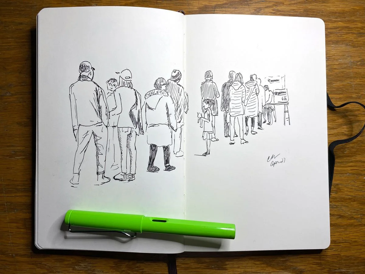 A sketch of people standing in line on an open notebook with a green pen resting on the bottom left, on a wooden table.