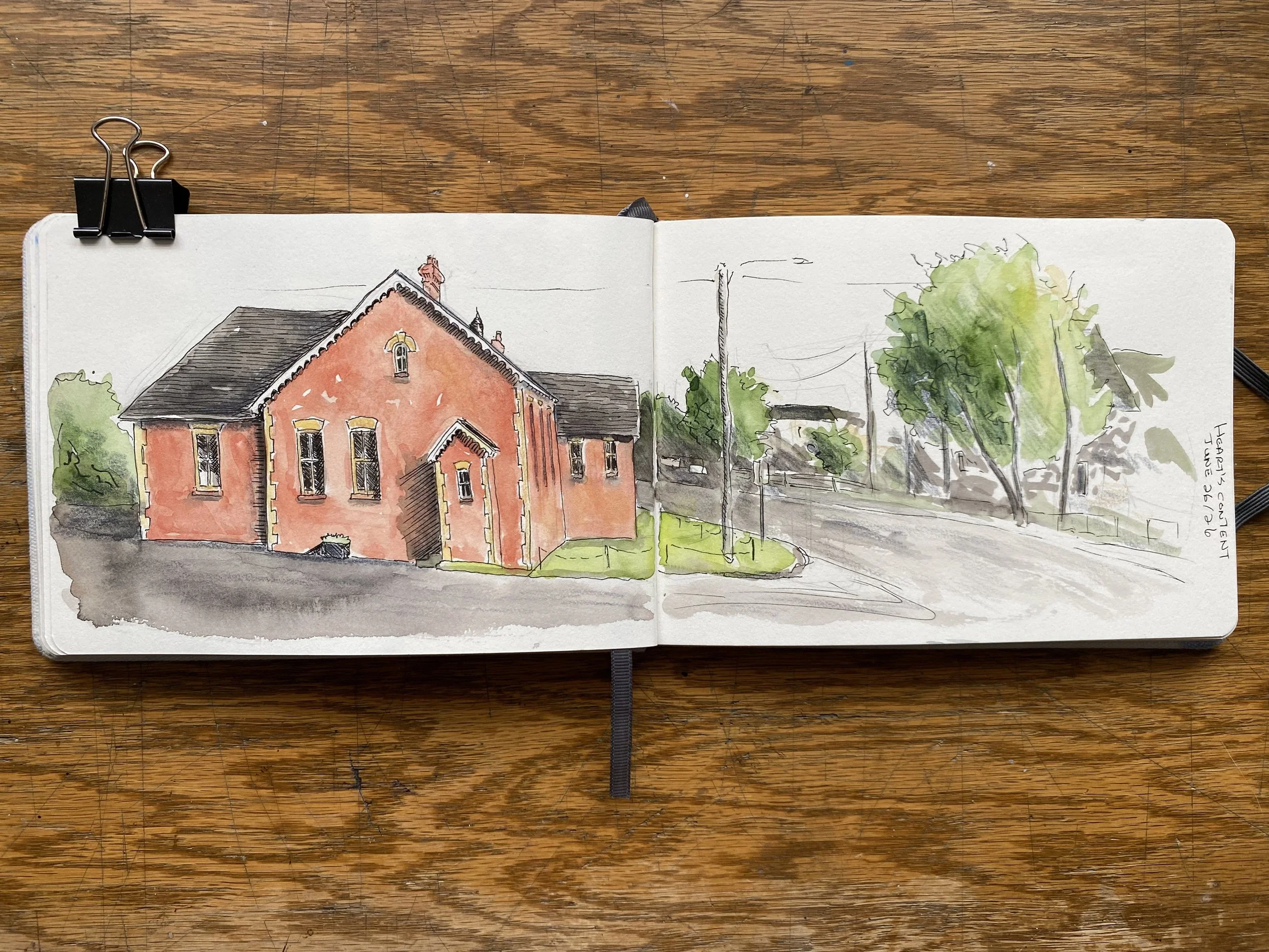 A watercolor sketch of a red brick house with a dark gray roof, surrounded by trees and a street with utility poles, on an open sketchbook placed on a wooden surface.
