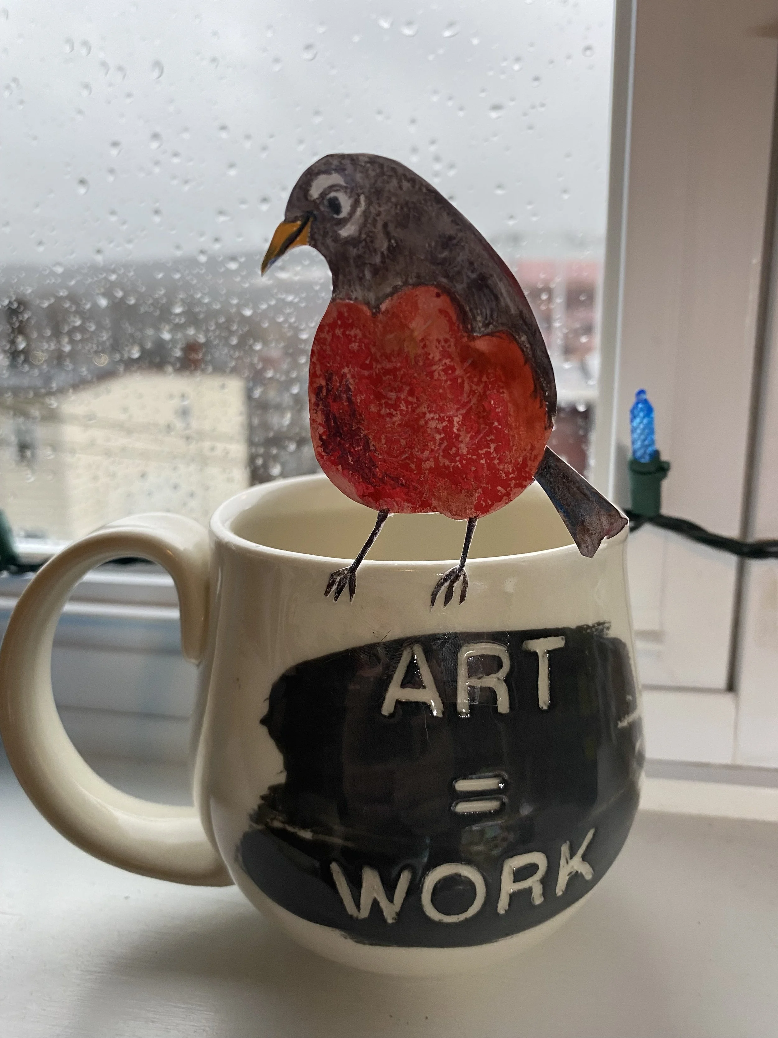 A coffee mug with the words 'HARD WORK' written on it, topped with a paper cutout of a bird with a red belly and a yellow beak, placed on a windowsill with raindrops on the window.
