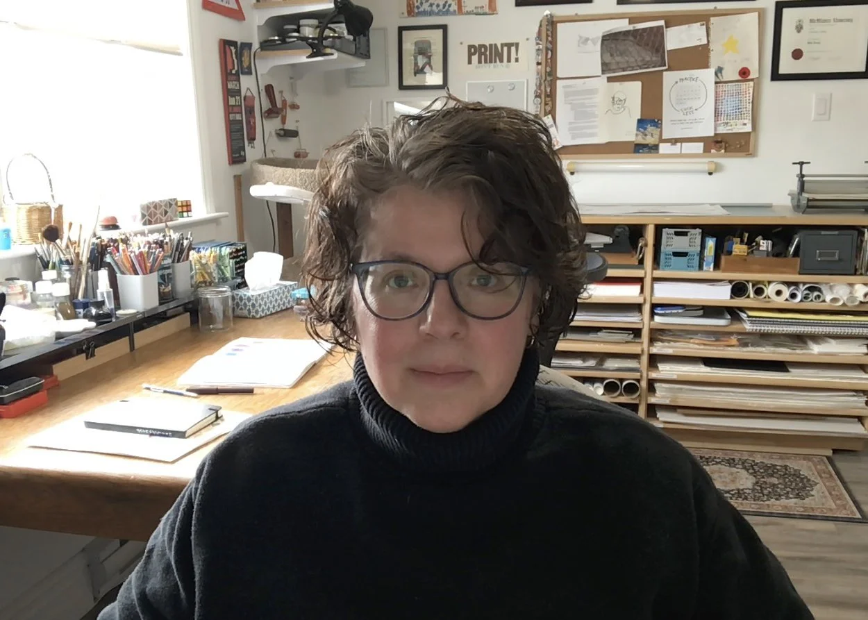 A woman with short, wavy brown hair and glasses sitting in a well-lit home office surrounded by art supplies, stacks of paper, and organizational shelves.