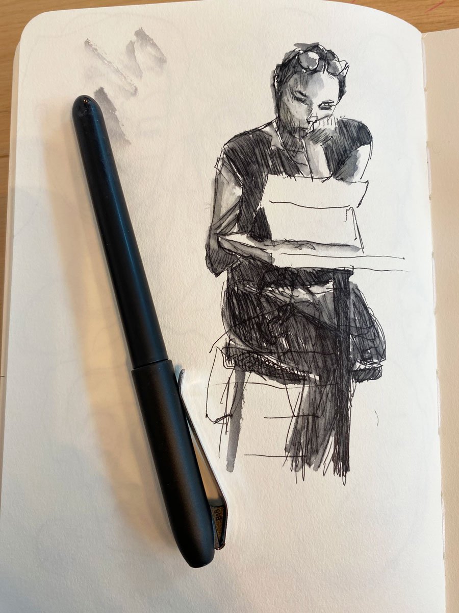 Sketch of a woman sitting at a table, reading or writing, with a pen placed on the paper in front of her. The woman has short dark hair, wears glasses on her head, and is dressed in a dark shirt.
