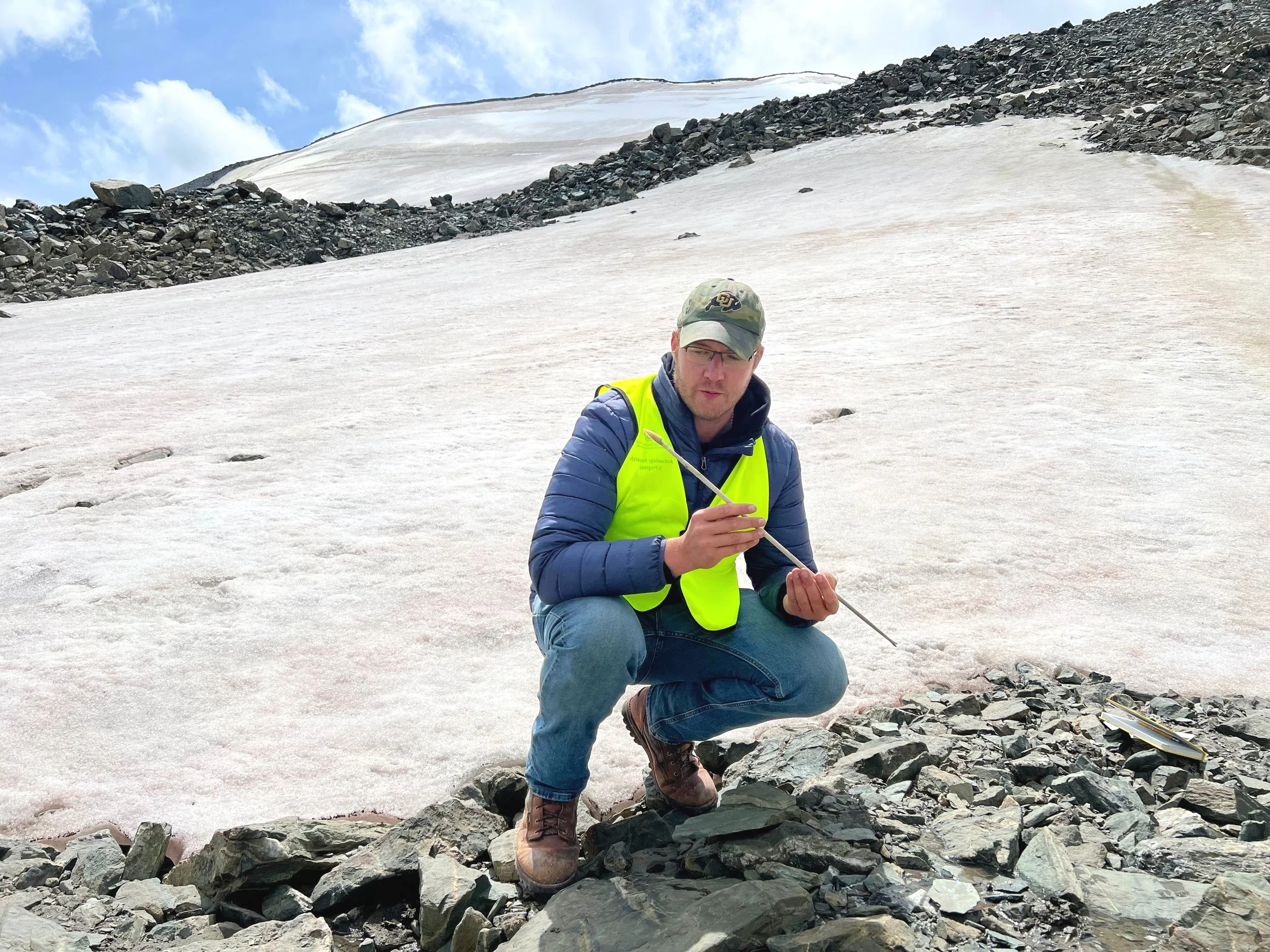 This image depicts the author recovering an ancient arrow from melting ice during archaeological fieldwork in the Altai Mountains of western Mongolia. He is squatting on rocks on top of a glacier holding the arrow in his hands.