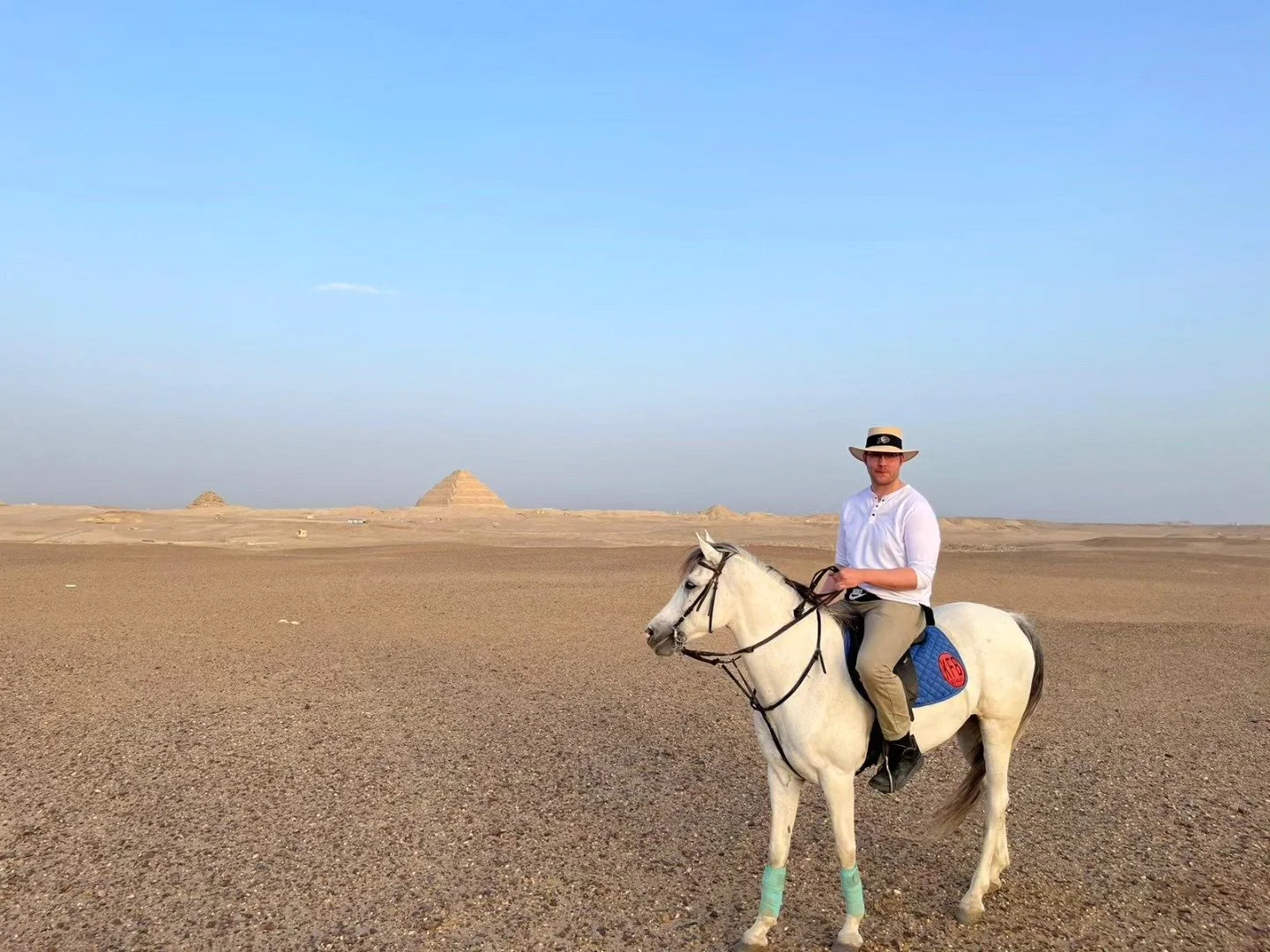 This image depicts the author at Saqqara, Egypt. He is on horseback in front of pyramids. He is wearing a white shirt and tan pants with a tan hat. His horse is white with dark grey mane and tail.