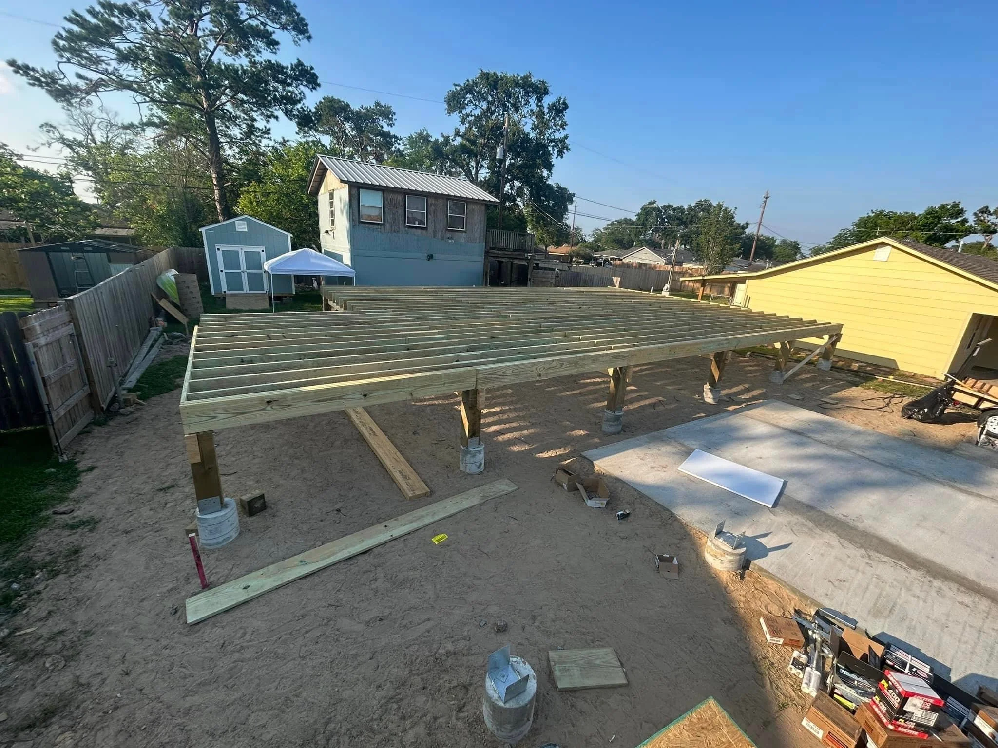 Under construction wooden deck or platform in backyard with fencing, sheds, and neighboring houses visible.