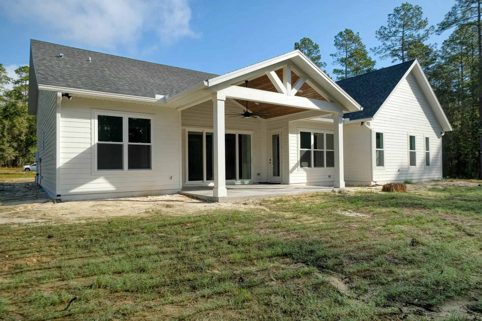 A newly built white house with a covered porch, large windows, and a high gable roof surrounded by a yard with grass and trees in the background.