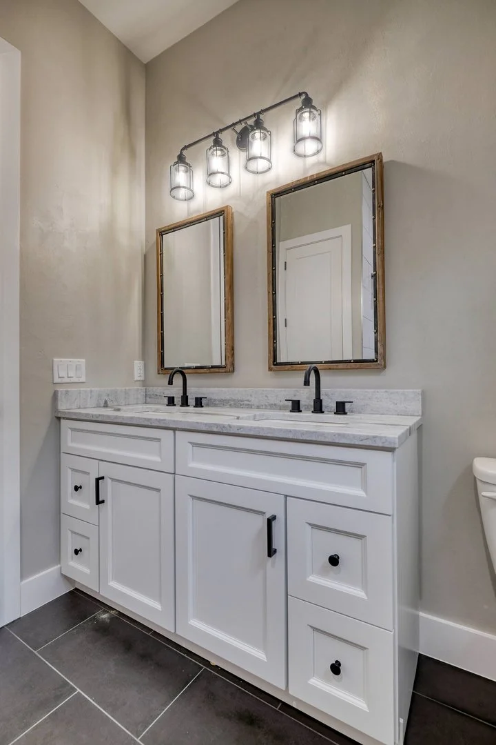 Modern bathroom vanity with white cabinets, marble countertop, two black faucets, two framed mirrors, and exposed bulb lighting fixture.