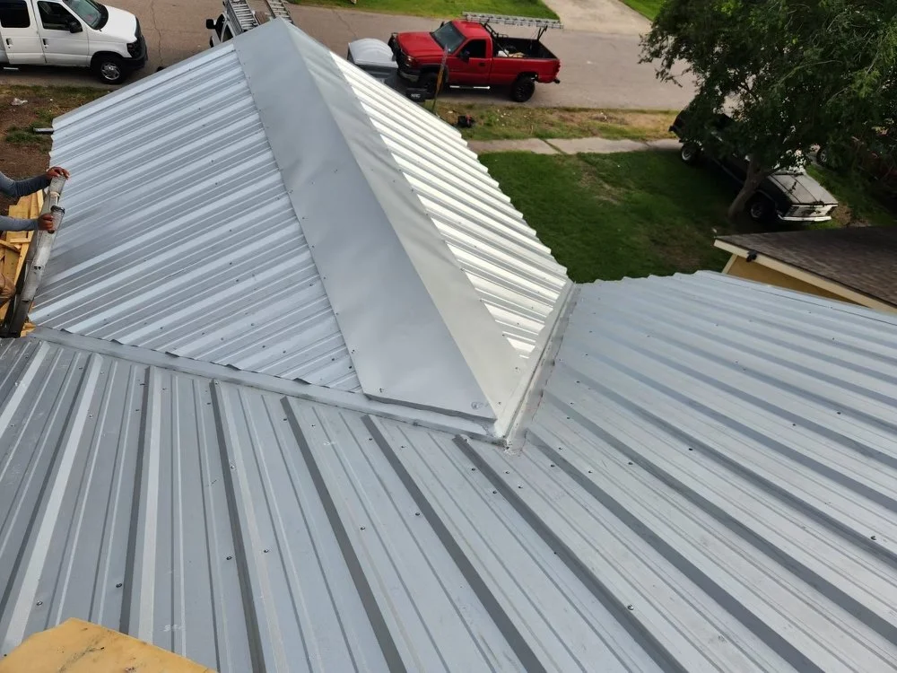 A close-up view of a metal roof with a worker installing a vent or chimney at the peak.